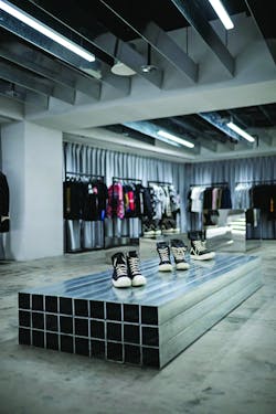Angled freestanding footwear and accessories display units are backlit. Suspended strip fixtures illuminate floor-mounted reflective displays and rack-hung garments.